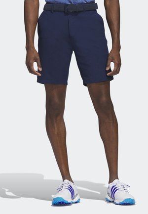 Mandlig model iført marineblå shorts med bælte, står barfodet på hvidt gulv, ben synlige fra talje til fødder.