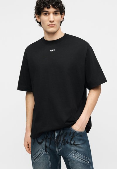 Νεαρός άντρας με σγουρά μαλλιά φοράει μια χαλαρή μαύρη μπλούζα με το κείμενο «OFF» και μπλε τζιν με μαύρο σχέδιο γραφής, στεκόμενος μπροστά σε μια απλή φόντο.