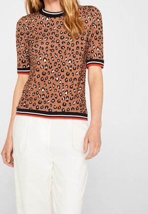 Kvinde iført en brun leopardmønstret top med korte ærmer og sorte, hvide og røde stribede manchetter og kant, kombineret med hvide bukser.