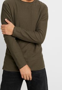 Maglietta a maniche lunghe verde oliva con texture a coste, scollo rotondo e maniche aderenti, indossata con jeans scuri.