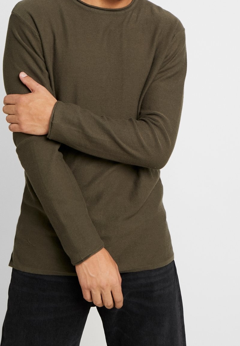 Maglietta a maniche lunghe verde oliva con texture a coste, scollo rotondo e maniche aderenti, indossata con jeans scuri.