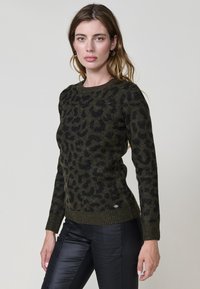 Olive zelena pulover s črnim leopardjim potiskom, narejen iz strukturejte pletenine, ima dolge rokave in okroglo ovratnico.