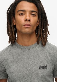 Grijze katoenen t-shirt met een ronde hals, korte mouwen en een klein geborduurd SuperDry-logo op de linkerkant van de borst. Glad textuur.