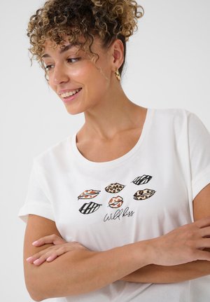 Lächelnde Frau mit lockigem Haar, die goldene Creolen und ein weißes T-Shirt mitTierdrucklippen und dem Text "Wild kiss" trägt.