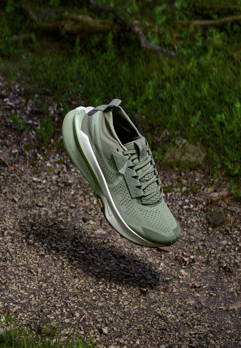 Groene sportieve schoen met een mesh bovenkant, geperforeerde details, een gevoerde zool en reflecterende accenten, opgehangen boven een grindoppervlak.
