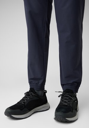 Legs wearing navy jogger pants and black low-top athletic sneakers with gray soles and patterned laces standing on a light gray floor.