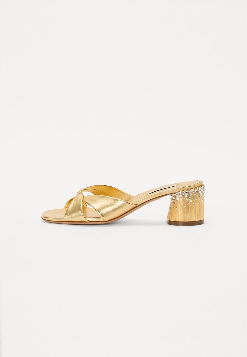Gold heeled sandal with crossed strap design, metallic finish, and a textured block heel adorned with silver rhinestones.