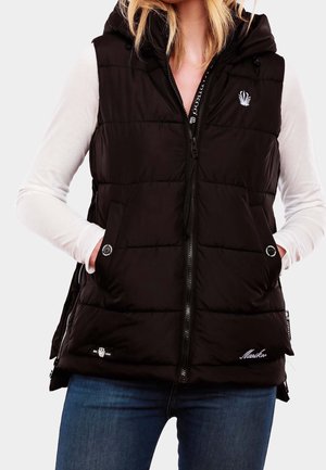 Veste noir matelassée sans manches avec capuche, fermeture éclair à l'avant et poches, portée sur un haut blanc à manches longues et un jean bleu.