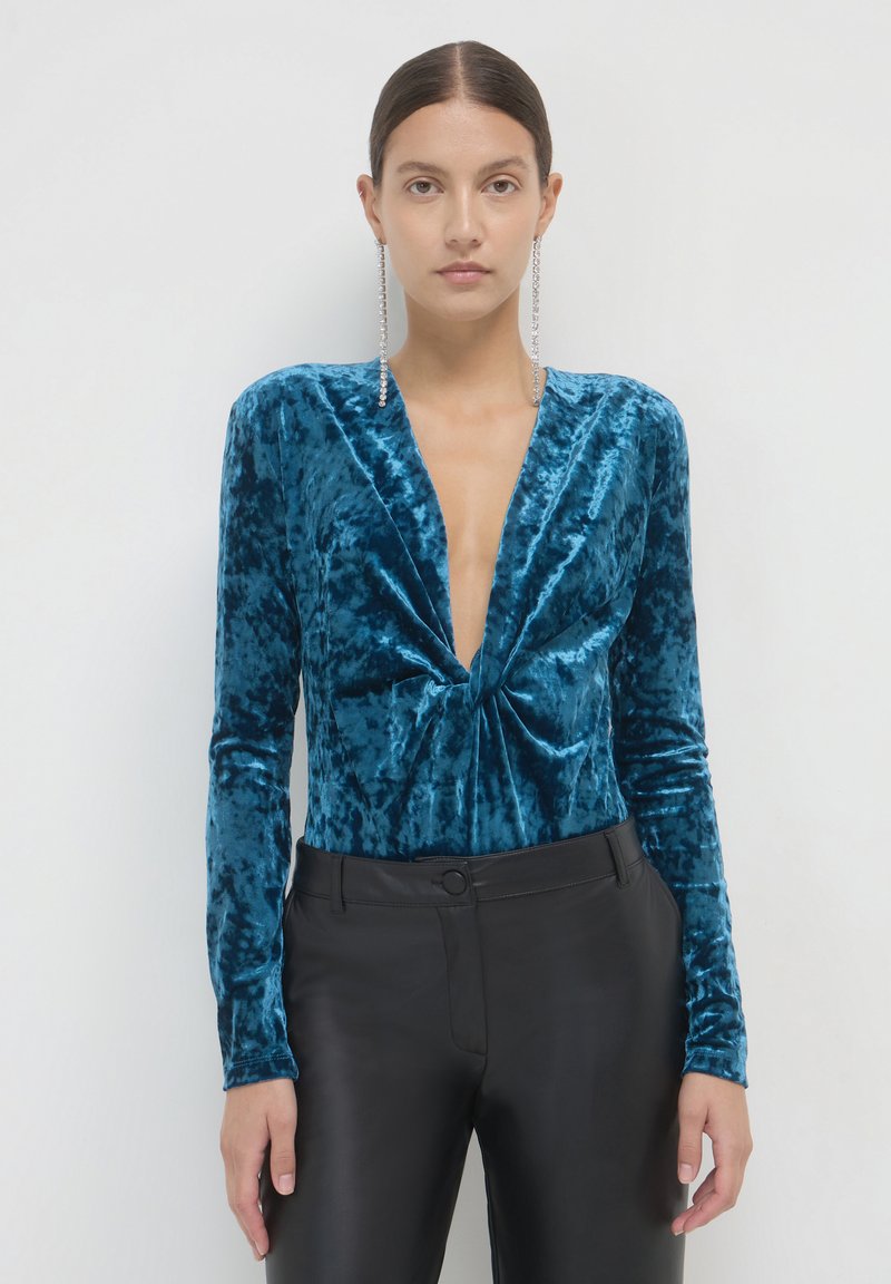 Body in velluto teal con maniche lunghe e scollatura a V profonda. Presenta un dettaglio frontale attorcigliato e si abbina a pantaloni neri a vita alta.