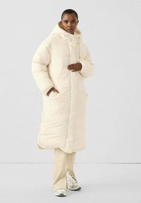 Roomwit oversized donzen jas met capuchon, steekzakken aan de voorkant en een trekkoord. Zachte, doorgestikte textuur, gecombineerd met lichtbeige broek en sneakers.