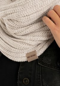 Gebreide stoffen sjaal in een zachte crème kleur met een textuurpatroon. Voorzien van een gestikte label met de tekst "KNIT FACTORY." Gedragen over een zwarte kledingstuk.