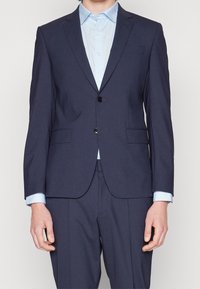 Marinblå skräddarsydd kostym med en knappstängning, två framfickor och en ljusblå skjorta under. Slät tyg och strukturerad passform.