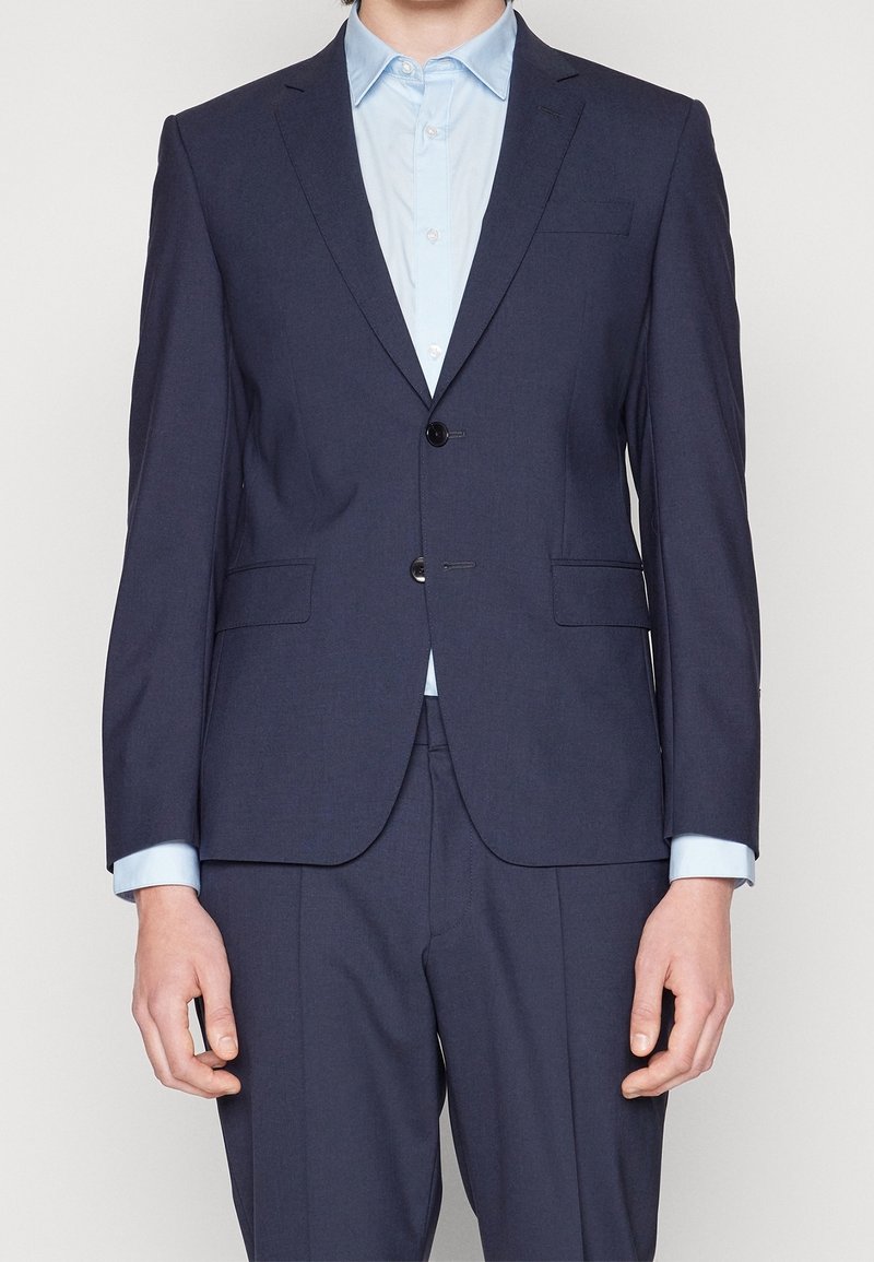 Marinblå skräddarsydd kostym med en knappstängning, två framfickor och en ljusblå skjorta under. Slät tyg och strukturerad passform.