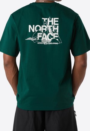 Osoba nosící tmavě zelené tričko s nápisem "The North Face" a bílou horskou grafikou na zádech, doplněné černými šortkami.