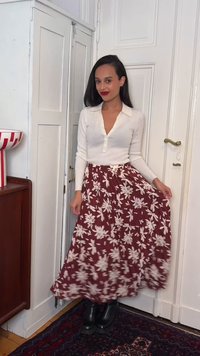 Γυναίκα που στέκεται μέσα, φοράει λευκό μπλουζάκι με μακριά μανίκια, μπορντό φλοράλ φούστα, μαύρες μπότες, κρατώντας την άκρη της φούστας, με ένα κόκκινο και λευκό μπολ σε έπιπλο κοντά.