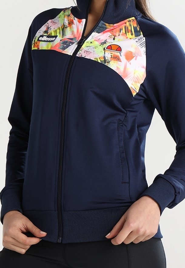 Femme portant une veste zippée bleu marine avec un imprimé abstrait coloré sur la poitrine et des logos sur les côtés gauche et droit.
