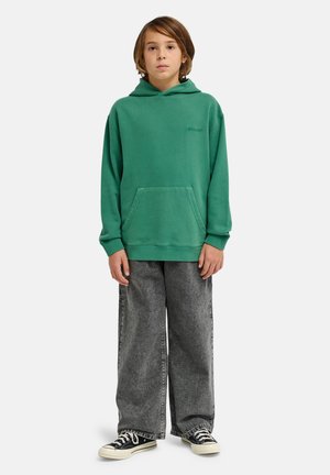 Enfant debout portant un sweat à capuche vert, un jean gris ample et des baskets noires basses sur un fond blanc.