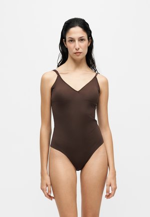 Vrouw met nat donker haar die een mouwloos bruin bodysuit draagt, staand tegen een effen lichtgrijze achtergrond.