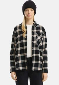 Jongeman met een zwarte beanie, een zwart en beige geruite overhemd over een witte top, en zwarte broek voor een effen achtergrond.