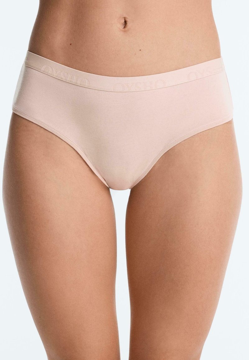 Buste féminin portant un slip beige clair sans couture taille mi-haute sur un fond blanc uni.