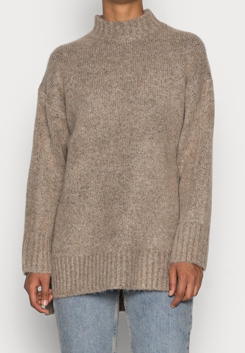 Beiger Strickpullover mit hohem, geripptem Kragen und langen Ärmeln. Verfügt über ein strukturiertes Muster und einen gerippten Saum. Kombiniert mit hellblauen Jeans.