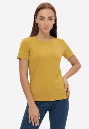 Giovane donna con capelli castani lisci indossa una maglietta giallo senape a maniche corte e jeans blu, in piedi con una mano sul fianco.