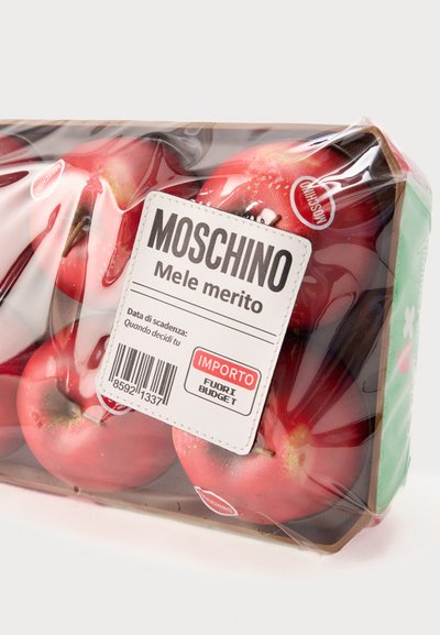 Συσκευασμένα κόκκινα μήλα με ετικέτα Moschino που αναγράφει "Mele merito", τυλιγμένα σε διάφανο πλαστικό με γραμμωτό κώδικα και προειδοποίηση προϋπολογισμού.