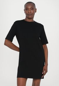 Czarna sukienka t-shirtowa z krótkimi rękawami, luźny krój, okrągły dekolt i subtelne logo na piersi. Gładki materiał o matowym wykończeniu.