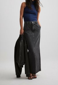 Falda maxi de mezclilla negra con cintura alta, combinada con una camiseta sin mangas azul marino con canalé. También incluye una chaqueta negra; el modelo lleva sandalias negras.