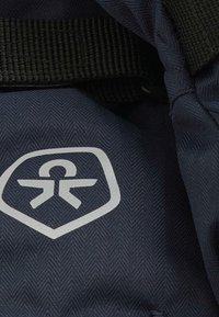 Unutrašnjost plave platnene torbe s herringbone teksturom, s bijelim logotipom u obliku štita i crnim remenom vidljivim u kutu.