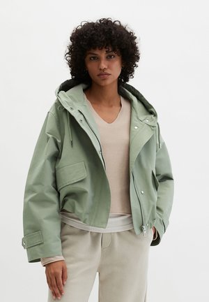Frau mit lockigem Haar, die eine hellgrüne Kapuzenjacke über einem beigen V-Ausschnitt-Pullover und beigen Hosen trägt, vor einem einfarbigen Hintergrund stehend.