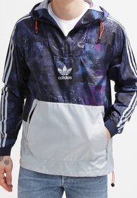 Muž má na sobě navy a šedou bundu Adidas s galaxií potiskem, bílé pruhy na rukávech a velkou přední kapsou, v kombinaci s modrými džíny.