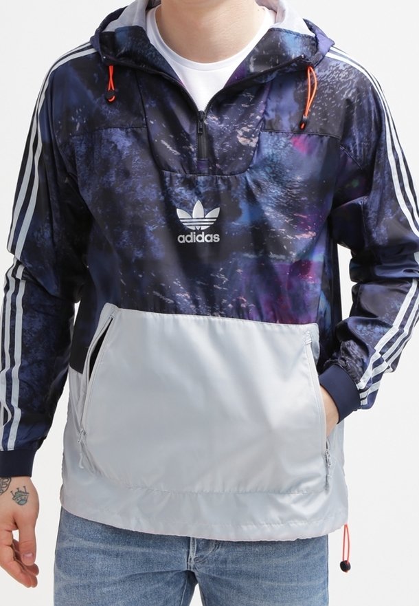 Muž má na sobě navy a šedou bundu Adidas s galaxií potiskem, bílé pruhy na rukávech a velkou přední kapsou, v kombinaci s modrými džíny.