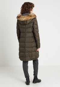 Cappotto imbottito verde oliva con rifinitura in pelliccia sintetica sul cappuccio, maniche lunghe e design trapuntato. Indossato sopra pantaloni grigio scuro e stivali neri.