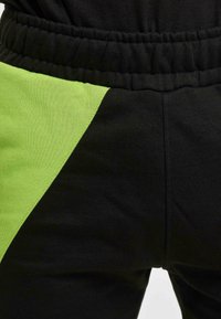 Melnas sporta bikses ar zaļu sānu paneli, kas aprīkotas ar elastīgu jostasvietu un mīkstu kokvilnas audumu. Dizainā ir ietverta kontrastējoša krāsu bloka elementa klātbūtne.