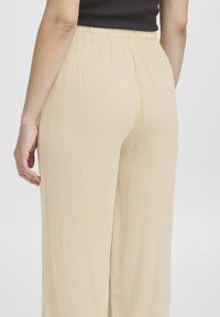 Beige geplooide broek met een lichtgewicht textuur. Elastische tailleband met subtiele verticale lijnen en een losse pasvorm. Achteraanzicht weergegeven.