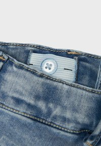 Pantalons en denim bleu clair avec une texture délavée, dotés d'un bouton bleu clair et d'une étiquette en denim. Détails de couture visibles.