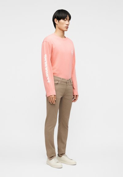 Νεαρός άνδρας που στέκεται στο πλάι, φορώντας ροζ πουκάμισο με μακριά μανίκια και λευκό κείμενο στα μανίκια, μπεζ παντελόνι και λευκά αθλητικά παπούτσια σε απλό φόντο.