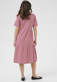 Robe rose à étages avec manches courtes, en tissu doux. Présente une jupe évasée et un design simple. Portée avec des chaussures plates noires.