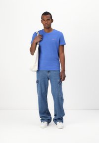 Blå bomull t-skjorte med korte ermer og en liten logo på brystet, kombinert med lyseblå cargo-jeans og hvite sneakers. Beigefarget totebag som henger over skulderen.