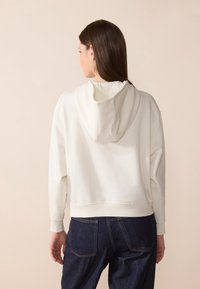 Sweatshirt blanc court avec une capuche, fabriqué en tissu doux, présentant une coupe décontractée et des poignets côtelés. Associé à un pantalon en denim bleu foncé.