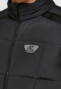 Schwarze Steppjacke mit hohem Kragen, ausgestattet mit einem Frontreißverschluss, gesteppter Struktur und einem Logoaufnäher auf der Brust.