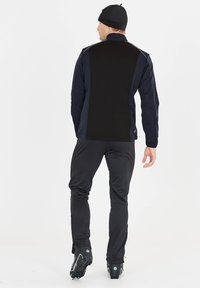 Veste de sport en bleu marine avec des accents noirs, dotée d'un col haut et de détails réfléchissants. Associée à un pantalon noir fuselé et des bottes noires.