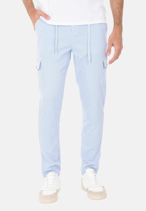 Uomo che indossa pantaloni cargo azzurri chiaro con tasche laterali e sneaker bianche, in piedi con una mano in tasca.