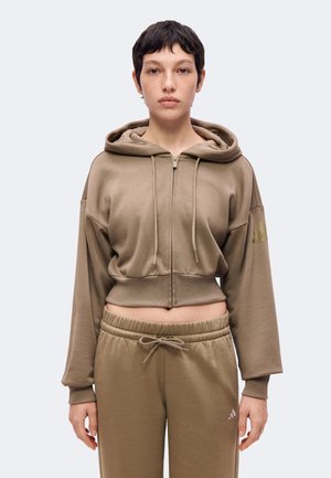 Mladá osoba s krátkymi tmavými vlasmi, ktorá má na sebe krátku hnedú mikinu na zips a k nej ladúce hnedé tepláky so šnúrkou.