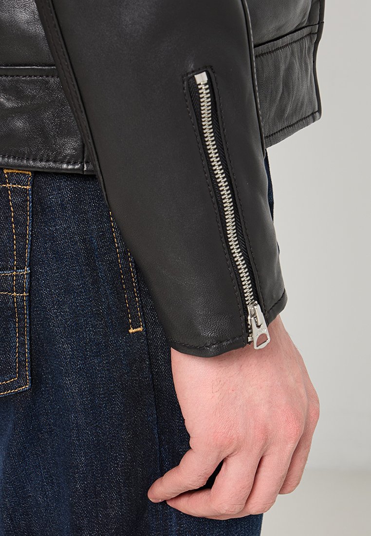 Manga de chaqueta de cuero negro con detalle de cremallera plateada. El primer plano muestra la textura suave, el ajuste entallado y un fondo de denim en contraste.