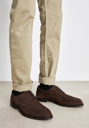 Jalat, joissa beigeihin käännetyt housut, mustat sukat ja tummanruskeat mokkanahkaiset nauhalliset juhlakengät, seisovat vaaleanharmaalla lattialla.