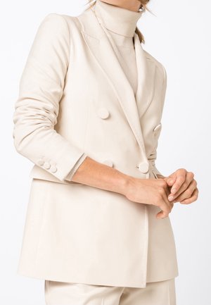 Kvinde iført en cremefarvet dobbeltradet blazer og en matchende rullekravesweater med hænderne foldet foran på en hvid baggrund.