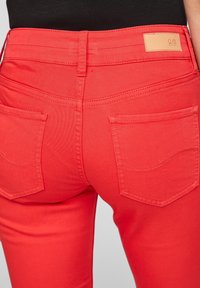 Punaiset denim-farkut, joissa on sileä pinta, keskikorkea vyötärö ja takataskut, joissa on hienovaraiset ompeluyksityiskohdat. Nahkainen etiketti vyötärön yläpuolella.
