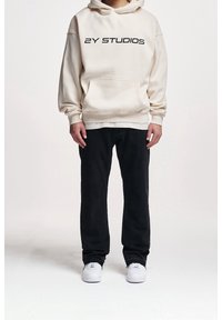 Kremowa bluza z kapturem z czarnym nadrukiem "2Y STUDIOS", przednia kieszeń, w zestawie z czarnymi dżinsami i białymi sneakersami. Prosty, casualowy design.